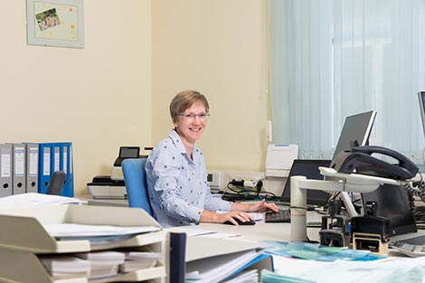 Frau Schabereiter sitzt am Bürotisch vor Ihrem Computer - im Hintergrund stehen Ordner.
